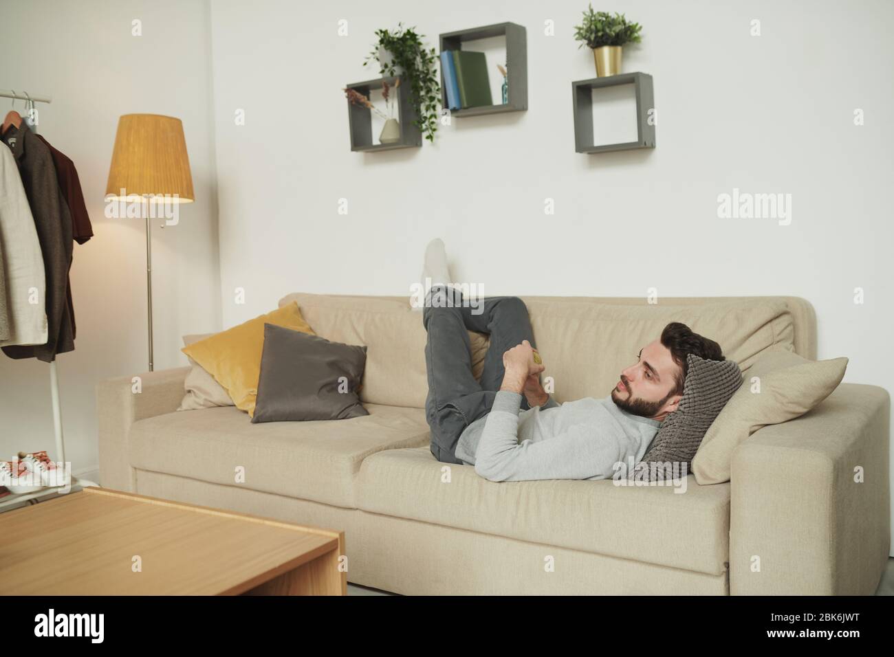Erholsamer Mann in Freizeitkleidung, der auf der Couch mit dem Kopf auf den Kissen vor dem fernseher liegt und Filme oder Sendungen im Wohnzimmer ansieht Stockfoto