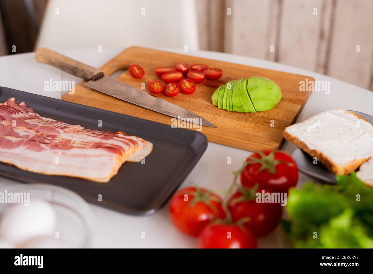 Speck, Avocado und Tomaten in Scheiben geschnitten auf Küchentisch in Prozess der Zubereitung gesundes Frühstück und zwei Sandwiches auf dem Teller in der Nähe Stockfoto