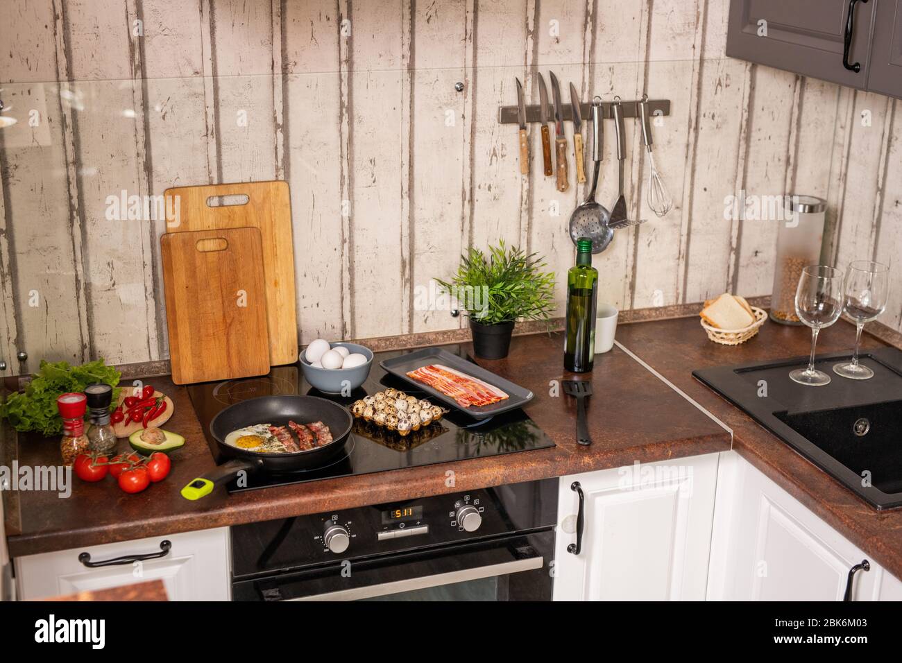 Teil der modernen Küche mit Spüle und Elektroherd mit Bratspeck und Eiern umgeben von Geschirr und Lebensmitteln Stockfoto
