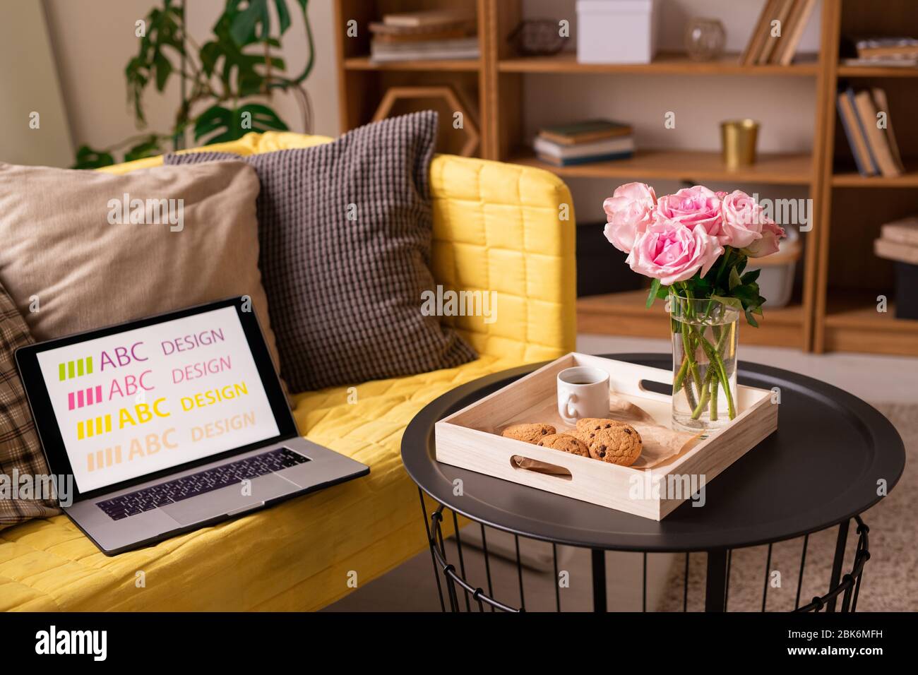 Kleiner runder schwarzer Tisch mit Rosen, Plätzchen und einer Tasse Kaffee, das gelbe Sofa mit Kissen und ein Laptop von Designer im Wohnzimmer Stockfoto