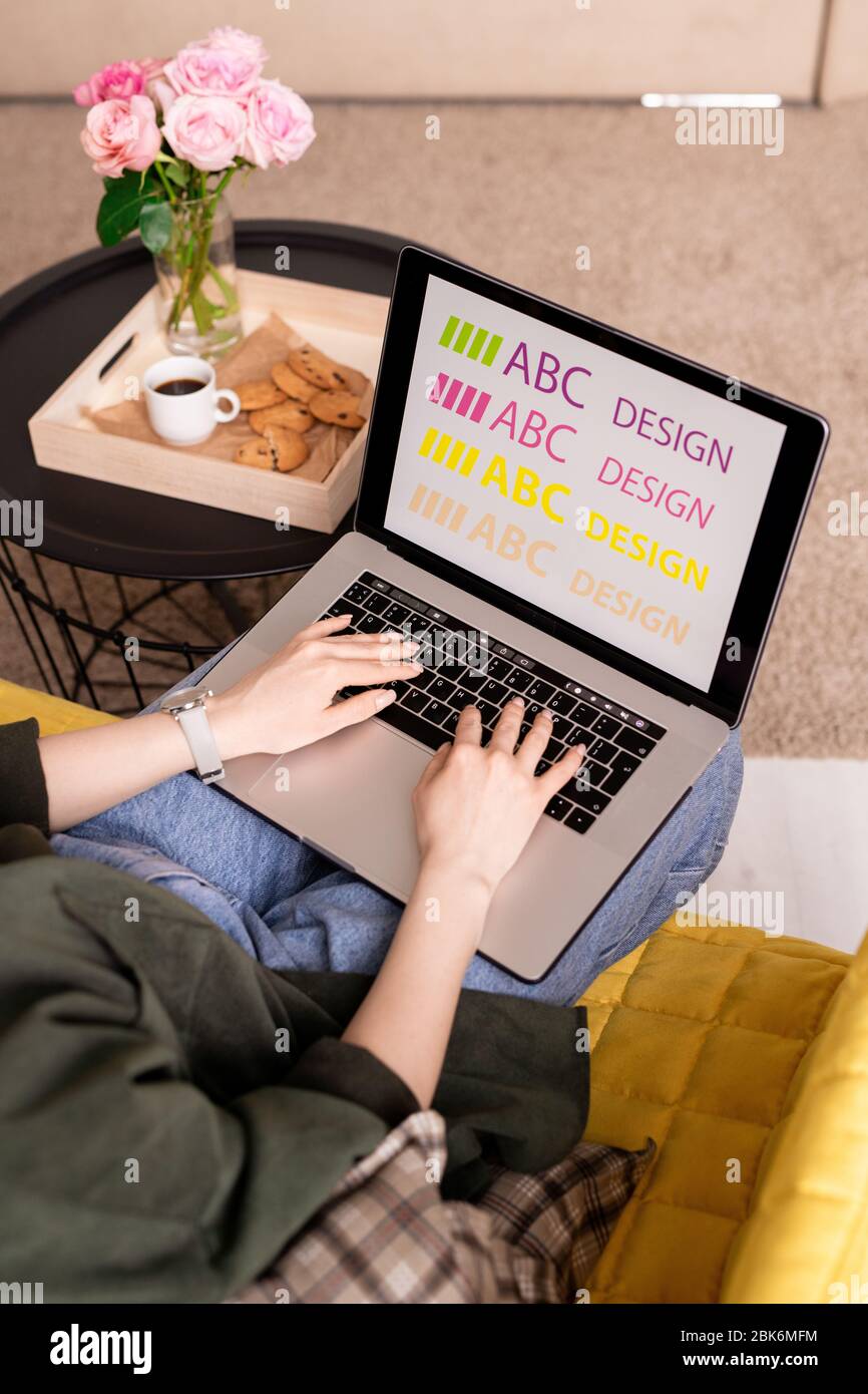 Hände der jungen Designerin auf Laptop-Tastatur während der kreativen Arbeit in der häuslichen Umgebung von kleinen Tisch mit Snack, Getränk und Rosen Stockfoto