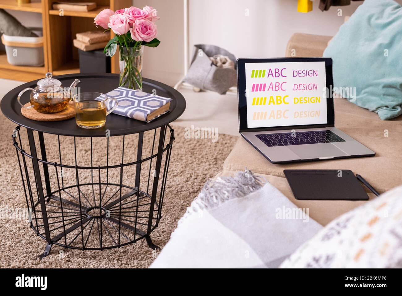 Kleiner Tisch mit Buch, grünem Tee und rosa Rosen auf weichem Teppich von bequemen Couch mit Pad, Stift und Laptop von Designer stehen Stockfoto