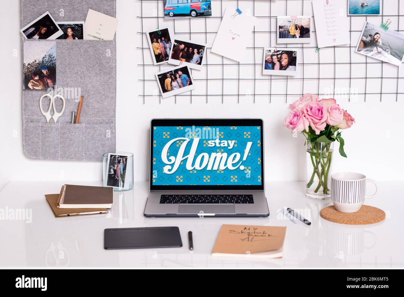 Wand mit Fotos und Schreibtisch mit Laptop, Bündel rosa Rosen, Pad mit Stift, Stifte, Notizbücher, Skizzenbuch und Becher mit Getränk Stockfoto