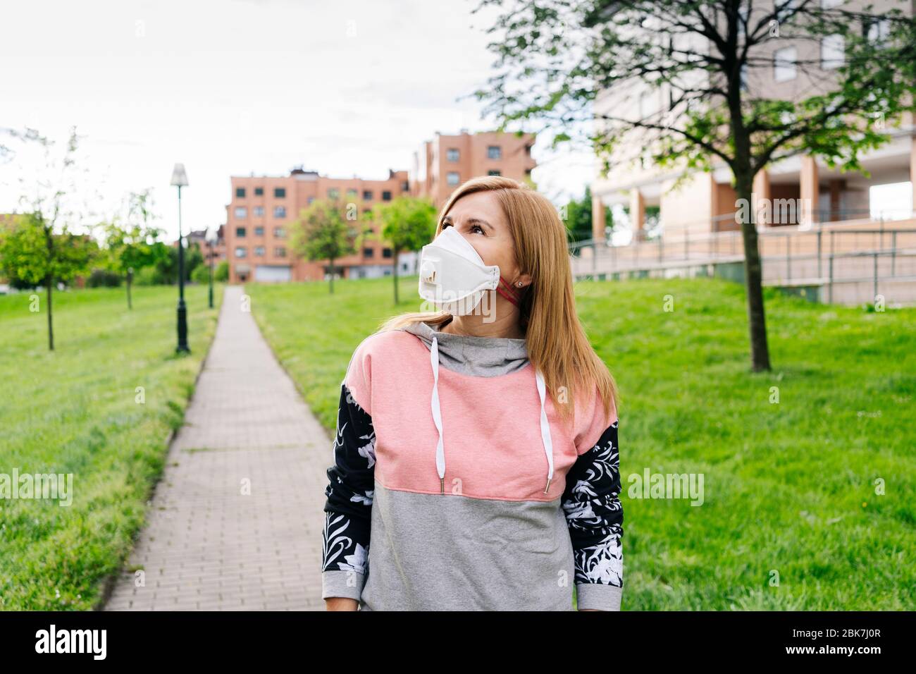Eine Frau mit einer Maske im Gesicht geht durch einen Park mit grüner Vegetation in einem Wohngebiet mit Gebäuden im Hintergrund Stockfoto