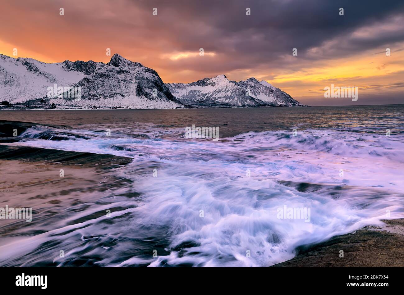 Blick über den Ersfjord von bunten Felsen bei Sonnenuntergang und Felsenbecken bis zu schneebedeckten Bergen an einem dunklen bewölkten Tag, Cape Tungeneset, Senja, Norwegen. Europa. Lang e Stockfoto