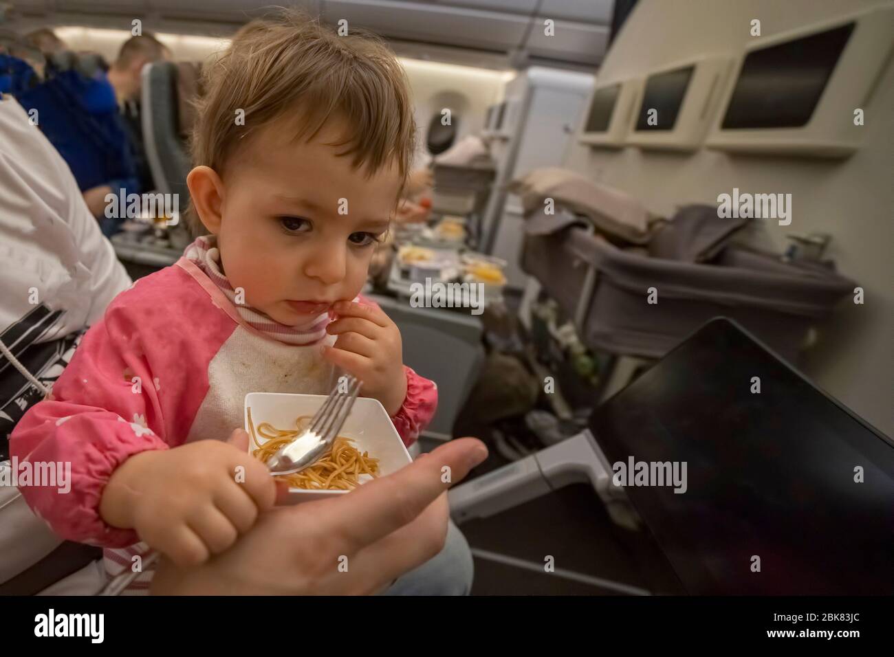Komfortabler Flug mit einem Kleinkind Konzept. Ein kleines niedliches hungriges Kind sitzt in einem Flugzeug auf dem Schoß seiner Eltern und isst leckeres Essen an Bord mit Stockfoto