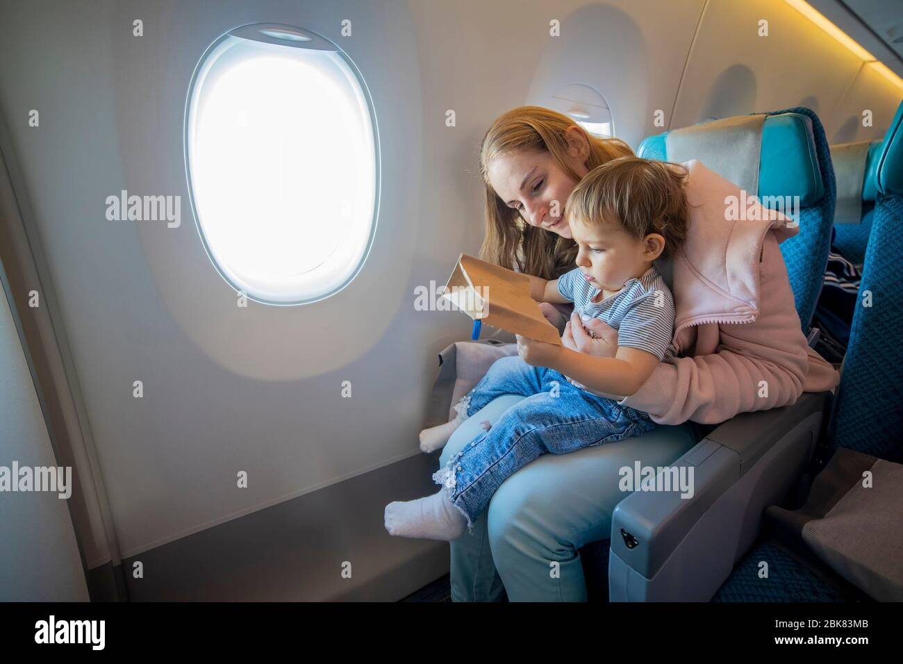 Junge schöne Mutter und kleines niedliches Kleinkind sitzen in einem Flugzeugsessel, öffnen Sie eine Papptasche mit Interesse. Stockfoto