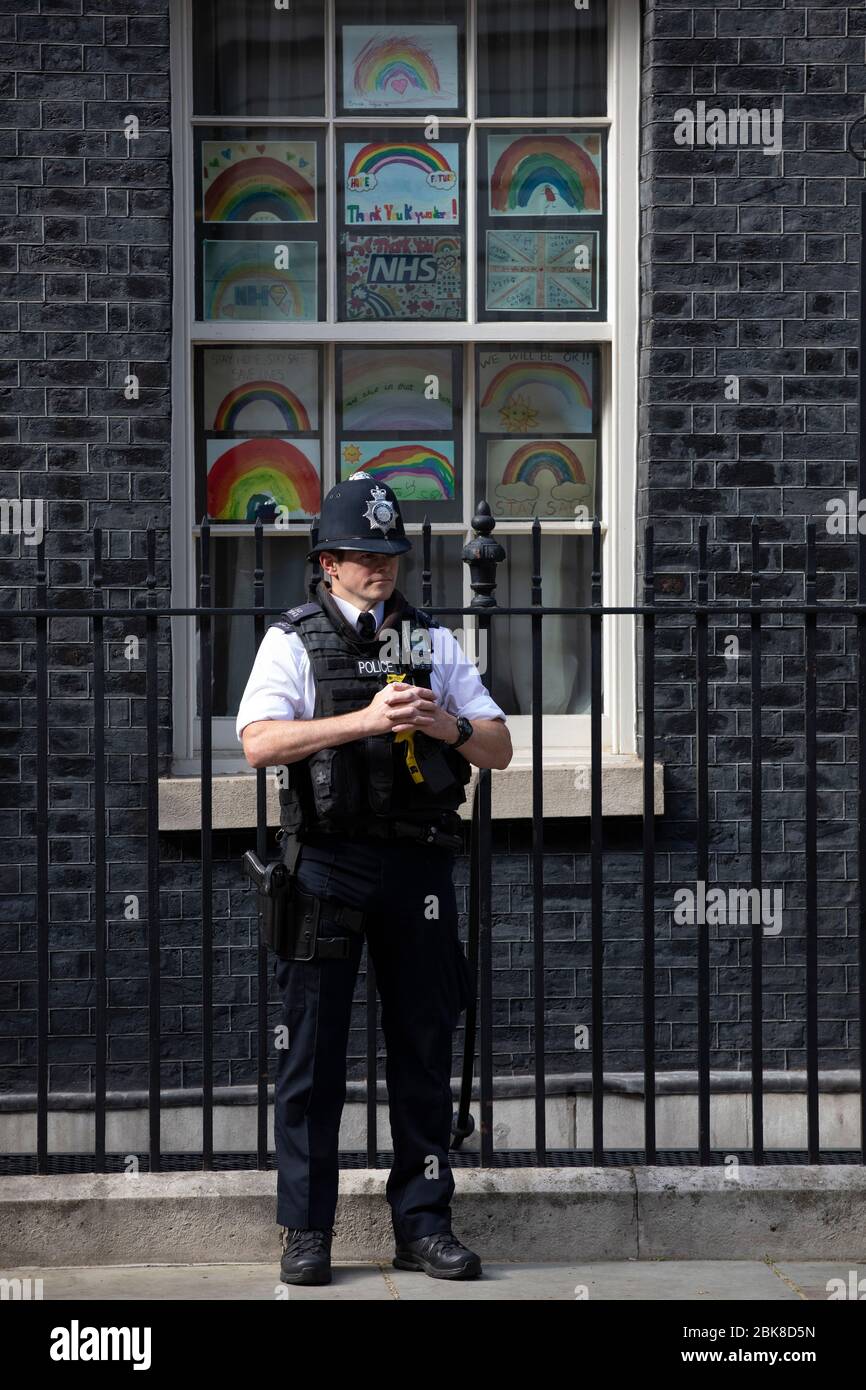 Ein Polizeibeamter steht vor "Regenbogenzeichnungen" in einem Fenster der Downing Street Nr. 10 in London, um NHS-Arbeiter gegen das Coronavirus in Großbritannien zu unterstützen Stockfoto