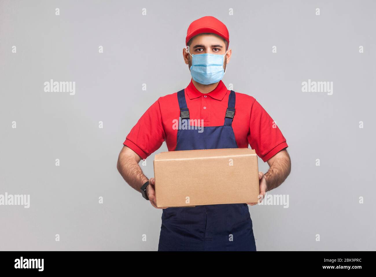 Lieferung in Quarantäne. Portrait des jungen Mannes mit chirurgischer medizinischer Maske in blauer Uniform und rotem T-Shirt, das steht und den Pappkarton am Gre hält Stockfoto
