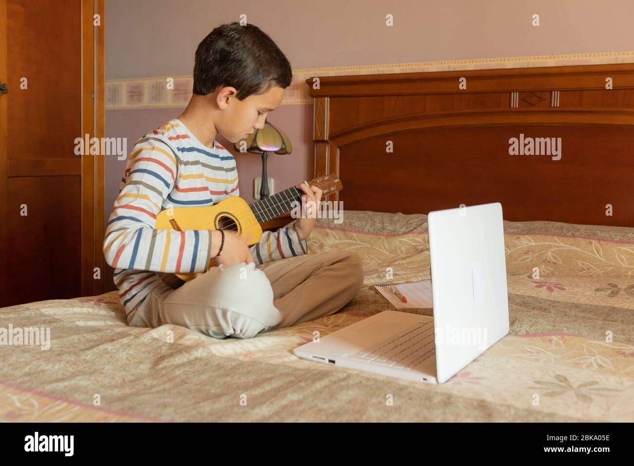 Junge, die einen Online-Kurs zu lernen, um die Timple spielen. Er sitzt zu Hause auf dem Bett und spielt Gitarre vor dem Laptop, während er ihm zuhört Stockfoto