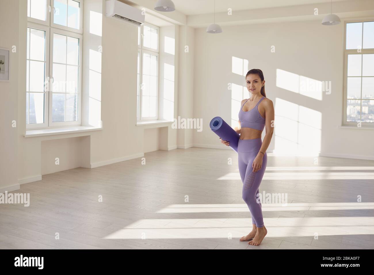 Fitness Yoga Sport Workout Frau. Eine glückliche Frau mit einer Yogamatte in den Händen steht in einem hellen Raum gegen eine weiße Wand. Stockfoto