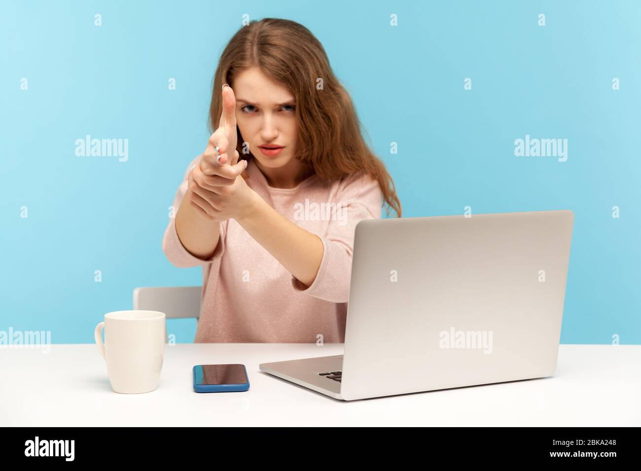 Wütende Frau Mitarbeiterin sitzt am Arbeitsplatz mit Laptop bedrohlich und zeigt mit der Pistole auf Kamera, schießen mit aggressivem Ausdruck, Targe zielen Stockfoto