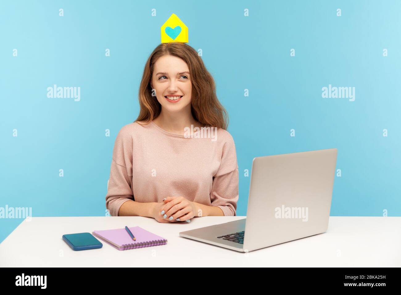 Junge freundliche positive Frau sitzt am Arbeitsplatz mit Laptop und halten kleine Papierhaus auf dem Kopf, träumen von eigenem Zuhause, Immobilienkauf. ind Stockfoto