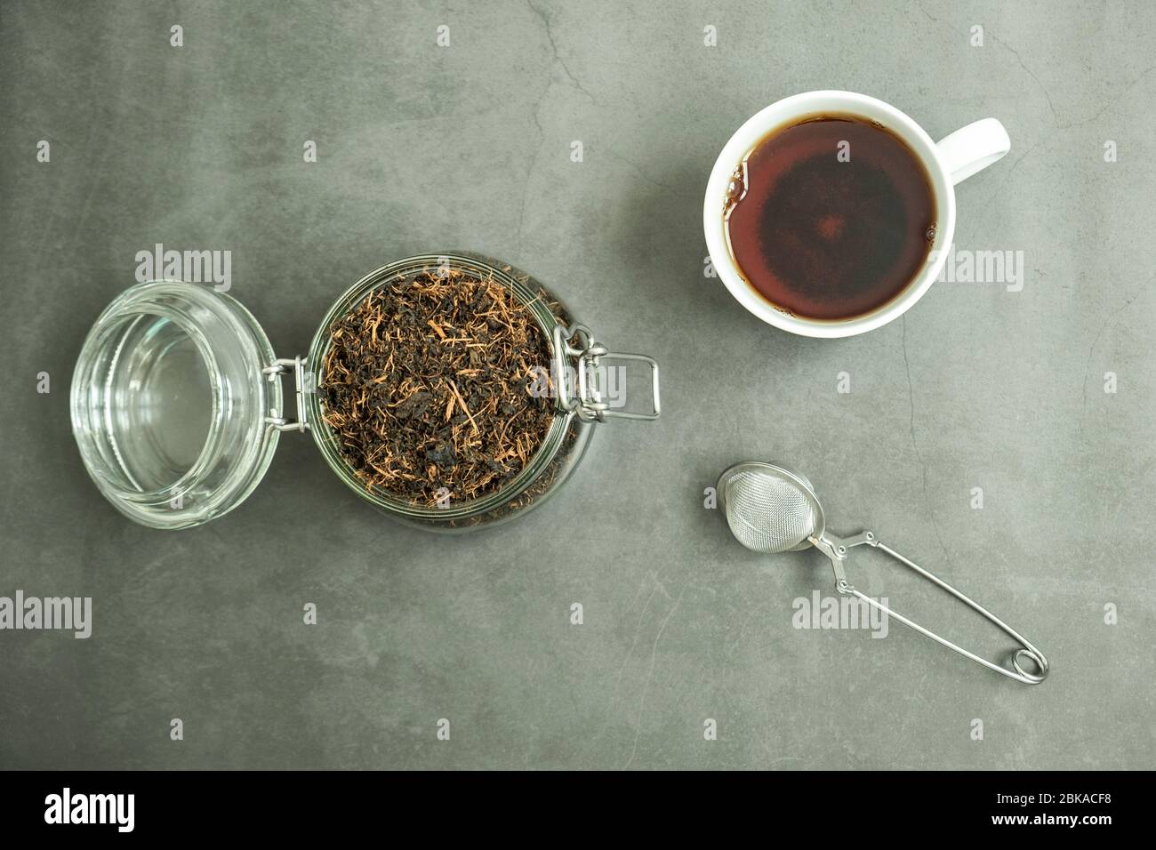 Trockenes schwarzes Teeblatt im Glasgefäß, Sieb und eine frische Tasse Tee auf Betongrund. Traditionelles, gesundes und biologisches Getränk. Draufsicht, flaches Lay Stockfoto