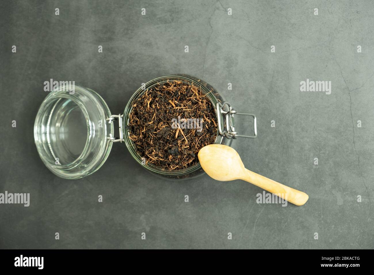 Trockenes schwarzes Teeblatt im Glasgefäß und einen Holzlöffel auf Beton, Steinhintergrund. Traditionelles, gesundes und biologisches Getränk. Draufsicht, flaches Lay. Stockfoto