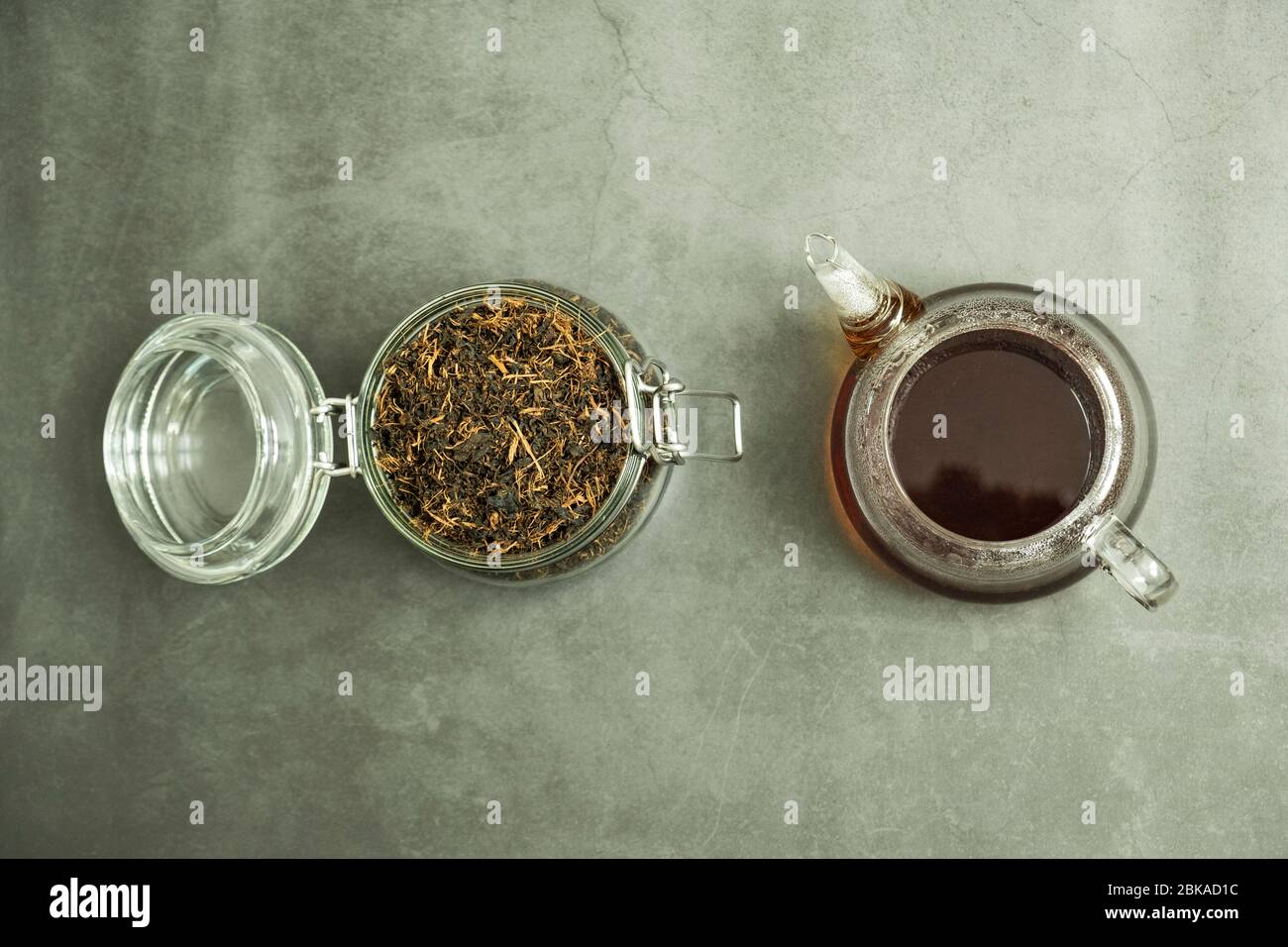 Trockenes schwarzes Teeblatt im Glasgefäß und frisch gebrühter frischer Tee in der Teekanne auf Beton, Steingrund. Traditionelles, gesundes und biologisches Getränk. Stockfoto