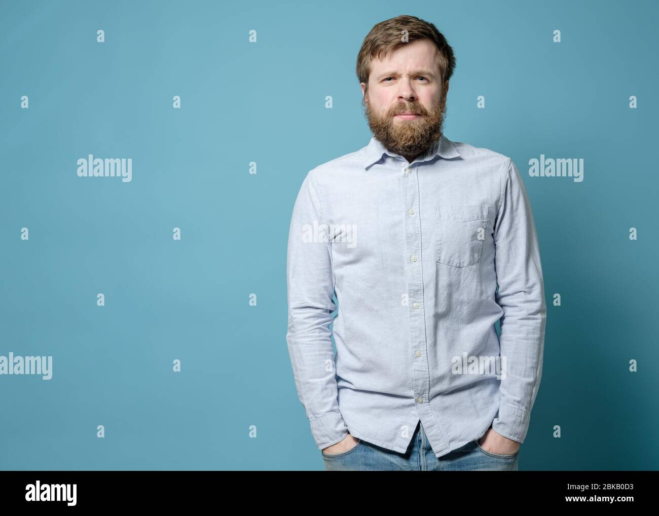 Alarmierte kaukasische Mann mit einem zotteligen Bart und einer überwucherten Frisur steht mit Händen in Jeans Taschen. Kopierbereich. Stockfoto