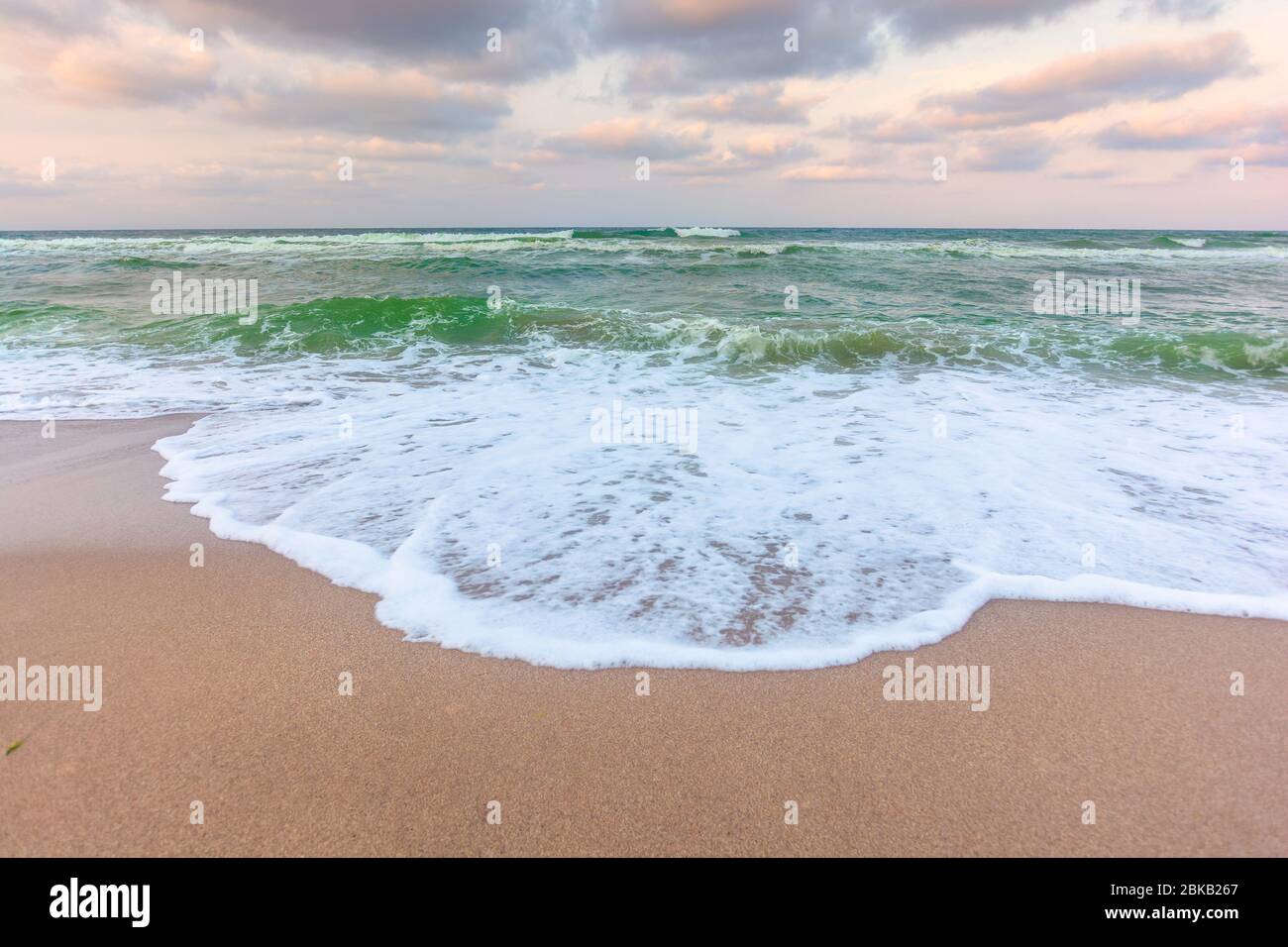 golden wolkigen Sonnenuntergang über dem grünen Meer Wasser. Wellen stürzt den Sandstrand. Klaren Horizont. Wechselnde windige Wetter Stockfoto