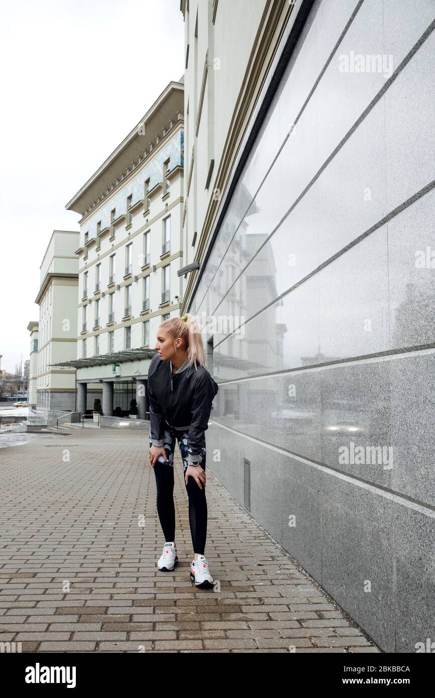 Hübsches blondes Mädchen mit Pferdeschwanz trainiert in Sportkleidung auf den Straßen einer Stadt. Sie trägt eine sportliche Jacke, Leggings und Sneaker. Stockfoto
