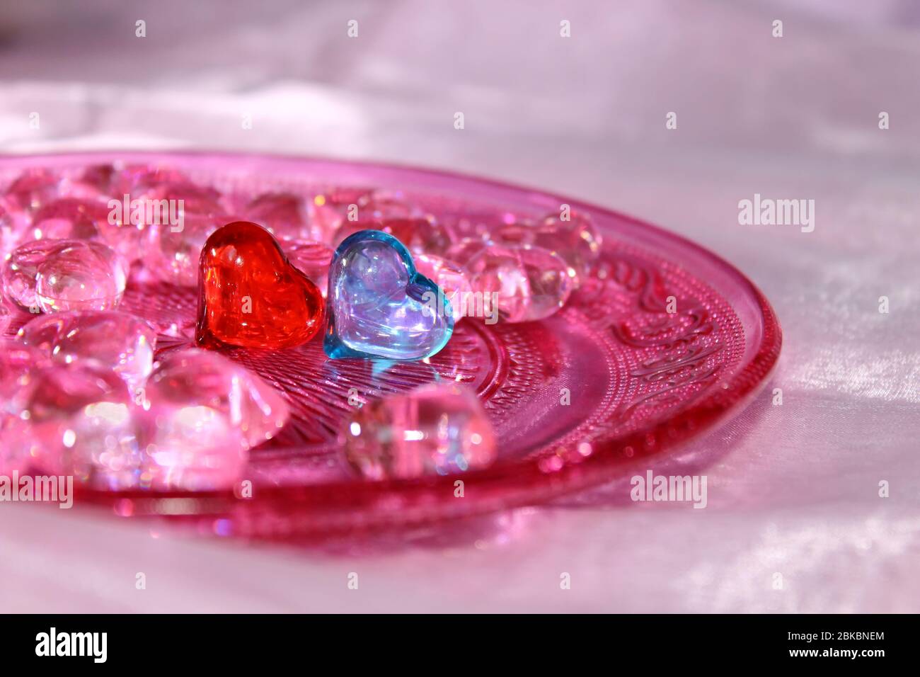 Ein rotes und ein blaues Glasherz, umgeben von rosa Herzen auf einer rosa Glasplatte mit Ornamentmuster. Hochzeitskarte Hintergrund. Stockfoto