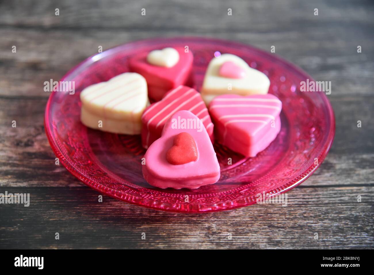 Herzförmige Pralinen in rosa, weiß und rot auf einem rosafarbenen Glasteller. Glasplatte hat ein Ornamentmuster. Dunkler Holztisch Hintergrund. Stockfoto