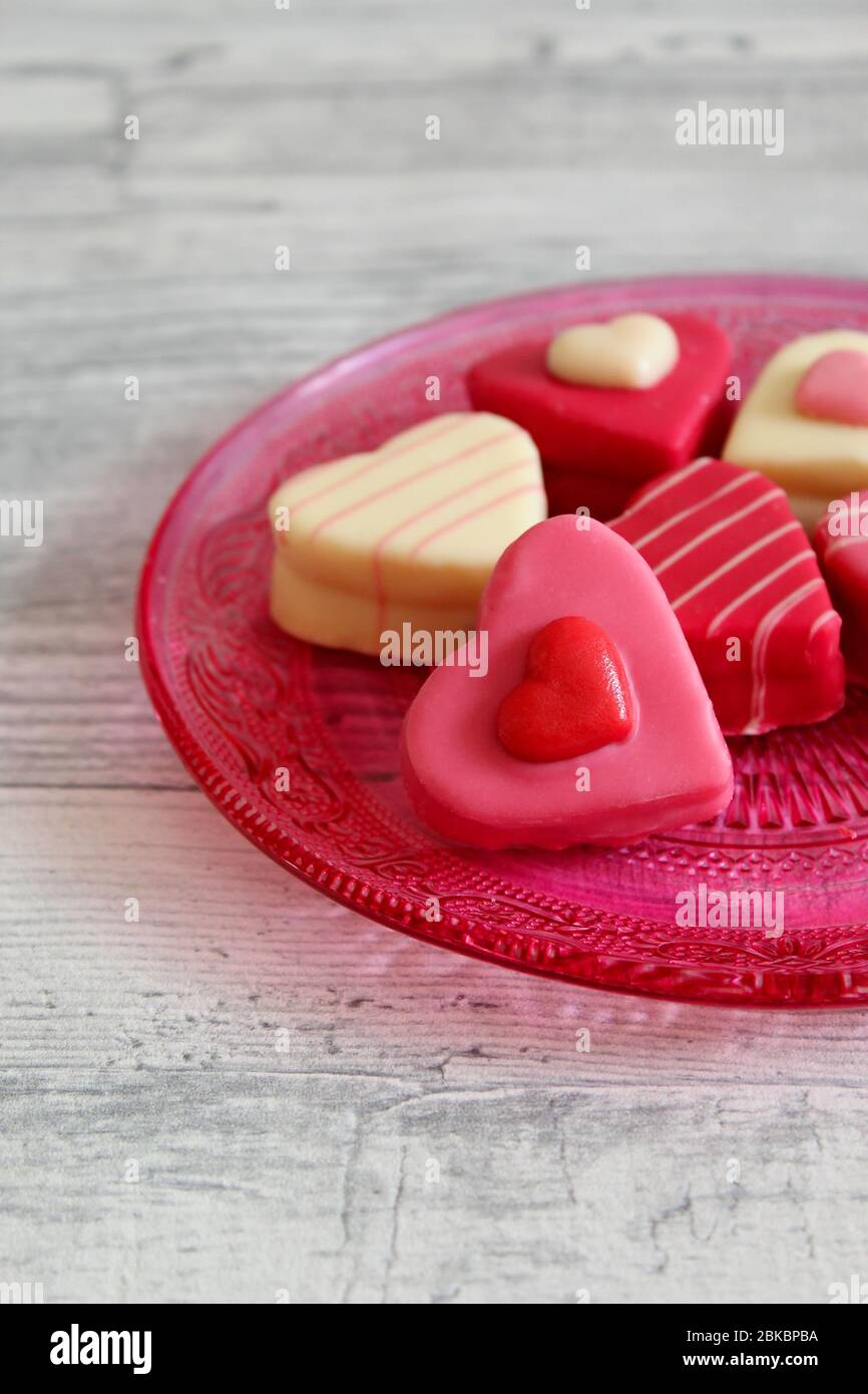 Herzförmige, bunte Pralinen (Petit Fours) auf einem rosa Glasteller mit Ornamentmuster. Heller Holztisch Hintergrund. Stockfoto