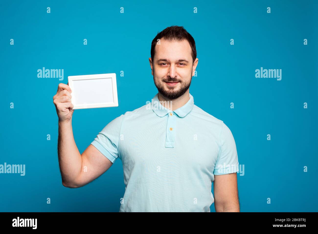 Ein Mann mit Bart auf blauem Hintergrund. Ein Mann hält einen weißen Rahmen. Stockfoto