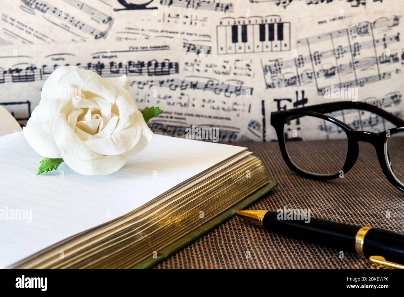 Weißes Papier Rose auf Notizbuch mit schwarzem und goldenem Stift gegen musikalische Notiztapete. Stockfoto