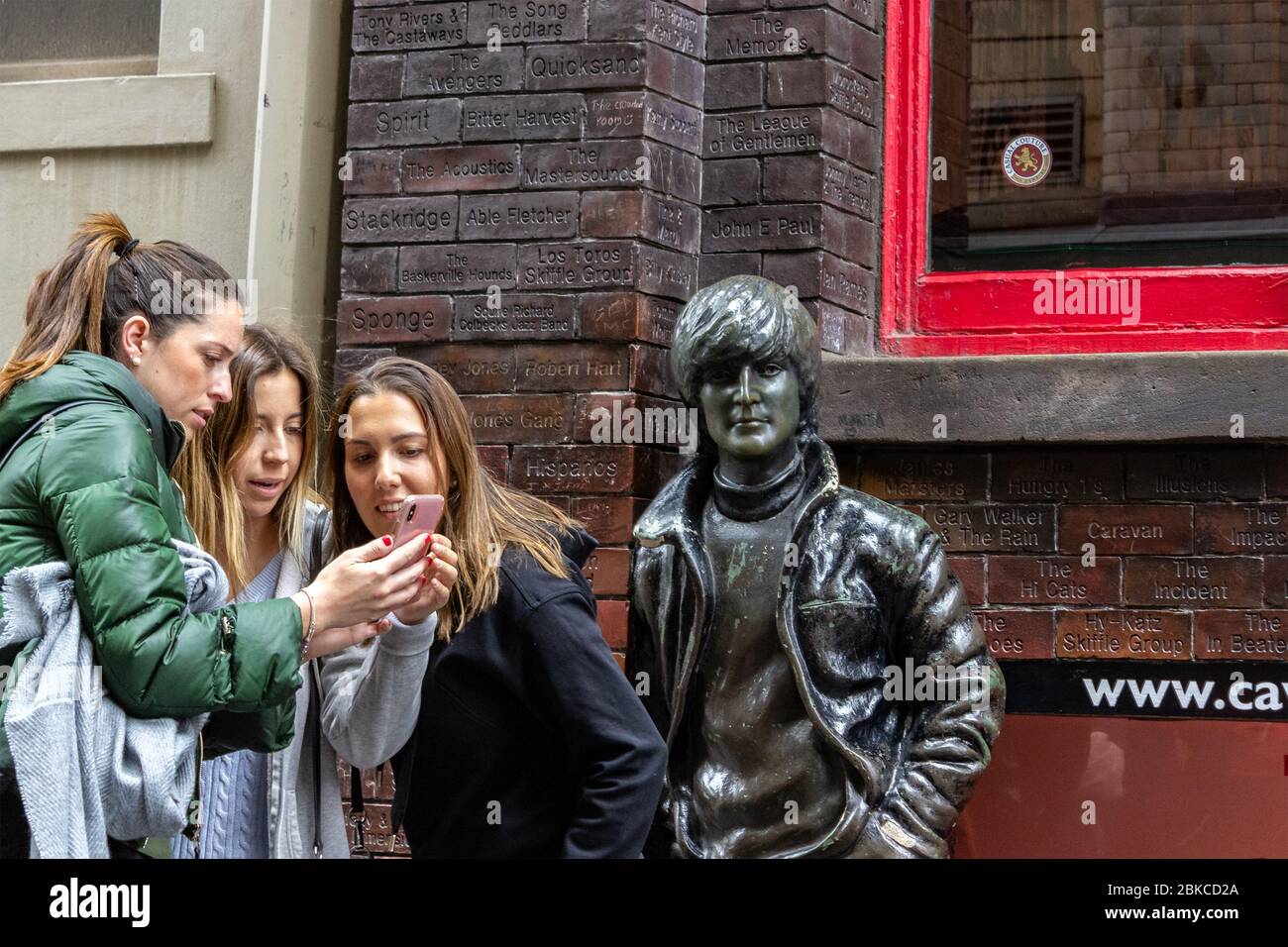 Freunde, die Fotos auf ihrem Smartphone neben der John Lennon Statue beim Besuch des Cavern Club, Mathew Street, ansehen, Stockfoto