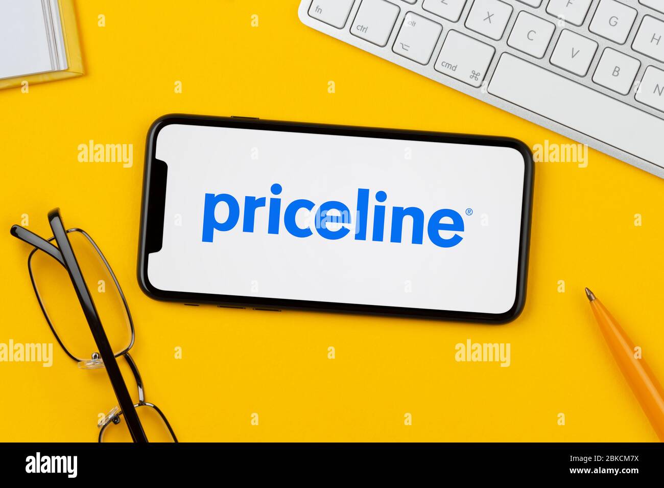 Ein Smartphone mit dem Priceline-Logo liegt auf einem gelben Hintergrund zusammen mit Tastatur, Brille, Stift und Buch (nur für redaktionelle Verwendung). Stockfoto