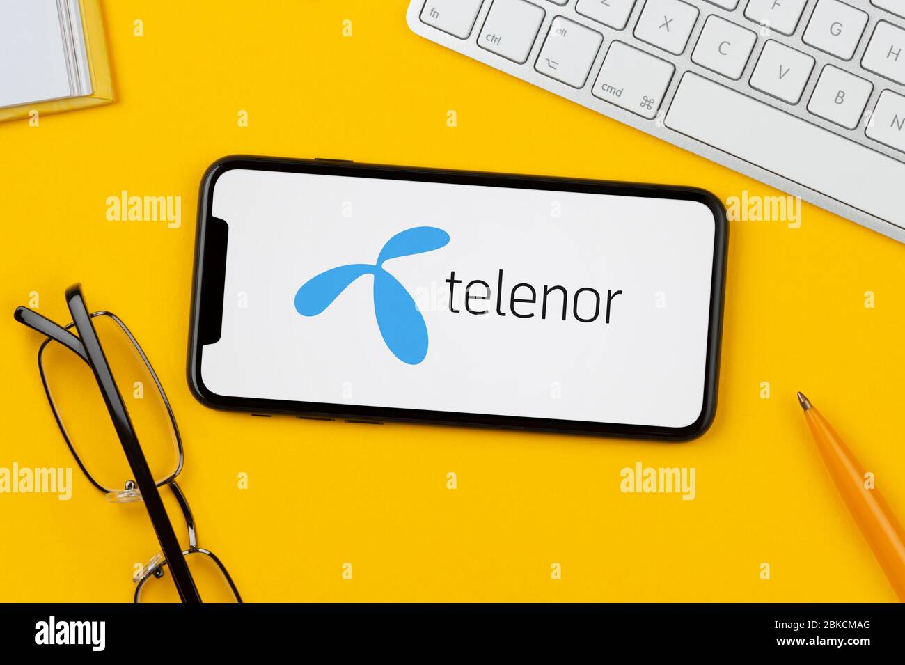 Ein Smartphone mit dem Telenor-Logo liegt auf einem gelben Hintergrund zusammen mit Tastatur, Brille, Stift und Buch (nur zur redaktionellen Verwendung). Stockfoto
