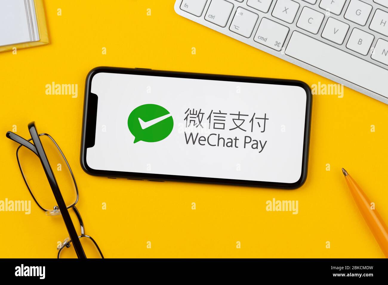 Ein Smartphone mit dem WeChat Pay-Logo liegt auf einem gelben Hintergrund zusammen mit Tastatur, Brille, Stift und Buch (nur für redaktionelle Verwendung). Stockfoto
