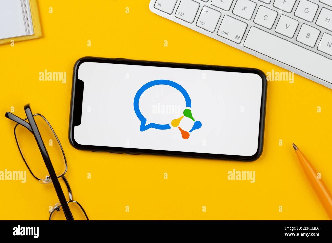 Ein Smartphone mit dem WeChat Work Logo liegt auf einem gelben Hintergrund zusammen mit Tastatur, Brille, Stift und Buch (nur redaktionelle Verwendung). Stockfoto