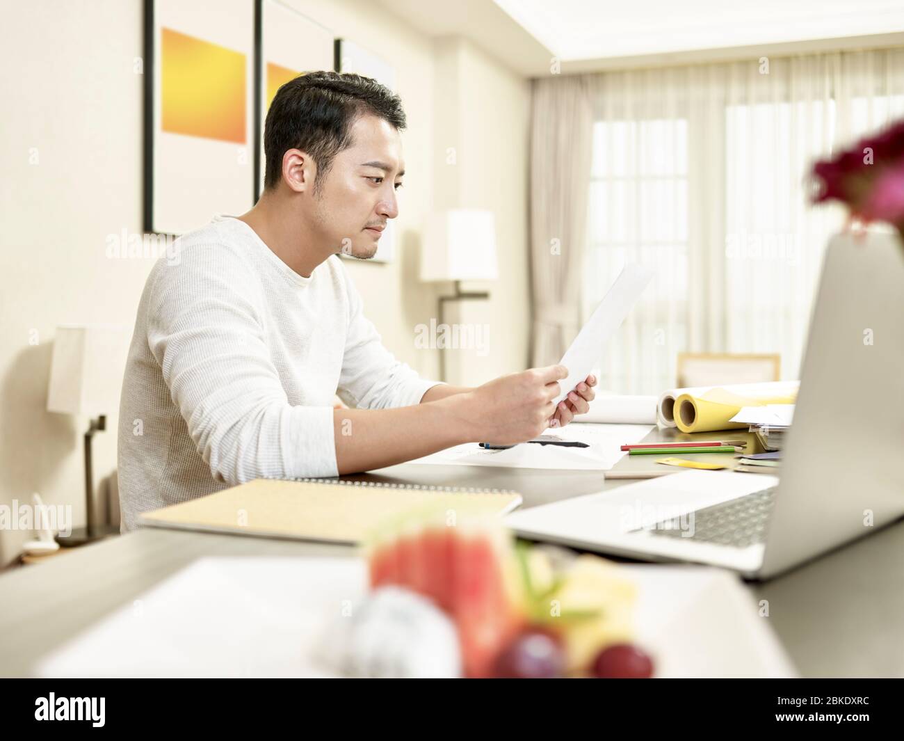 Junge asiatische Mann Design professionelle Arbeit von zu Hause aus sitzen an der Küche Theke Lesen eines Dokuments (Kunstwerk im Hintergrund digital verändert) Stockfoto