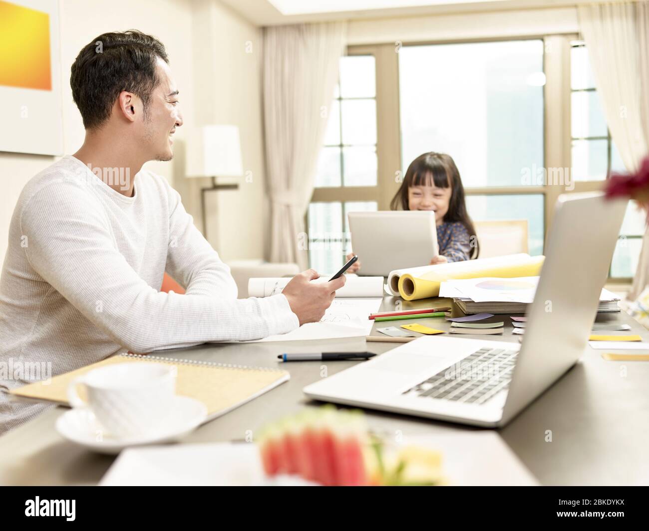 Junge asiatische Geschäftsmann kümmert sich um Kind während der Arbeit von zu Hause aus (Kunstwerk im Hintergrund digital verändert) Stockfoto
