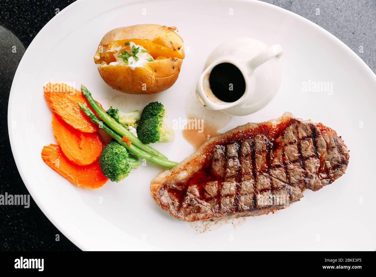 Draufsicht auf Holzkohle gegrilltes, knochenloses, trocken gereiftetes Top Loin Steak mit BBQ-Sauce, gebratenem Gemüse und gebackenen Kartoffeln auf einem weißen Teller. Stockfoto