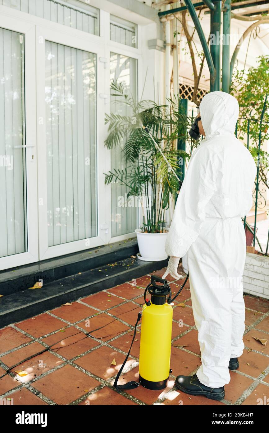 Techniker in Gefahrgutanzug Spritzen Chemikalien an Wänden und Fenstern des Hauses, um die Ausbreitung von Viren zu verhindern Stockfoto