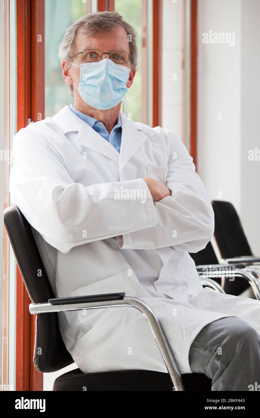 Arzt mit chirurgischer Maske sitzt in einem quirenden Raum und schaut selbstbewusst auf die Kamera - Fokus auf das Gesicht Stockfoto