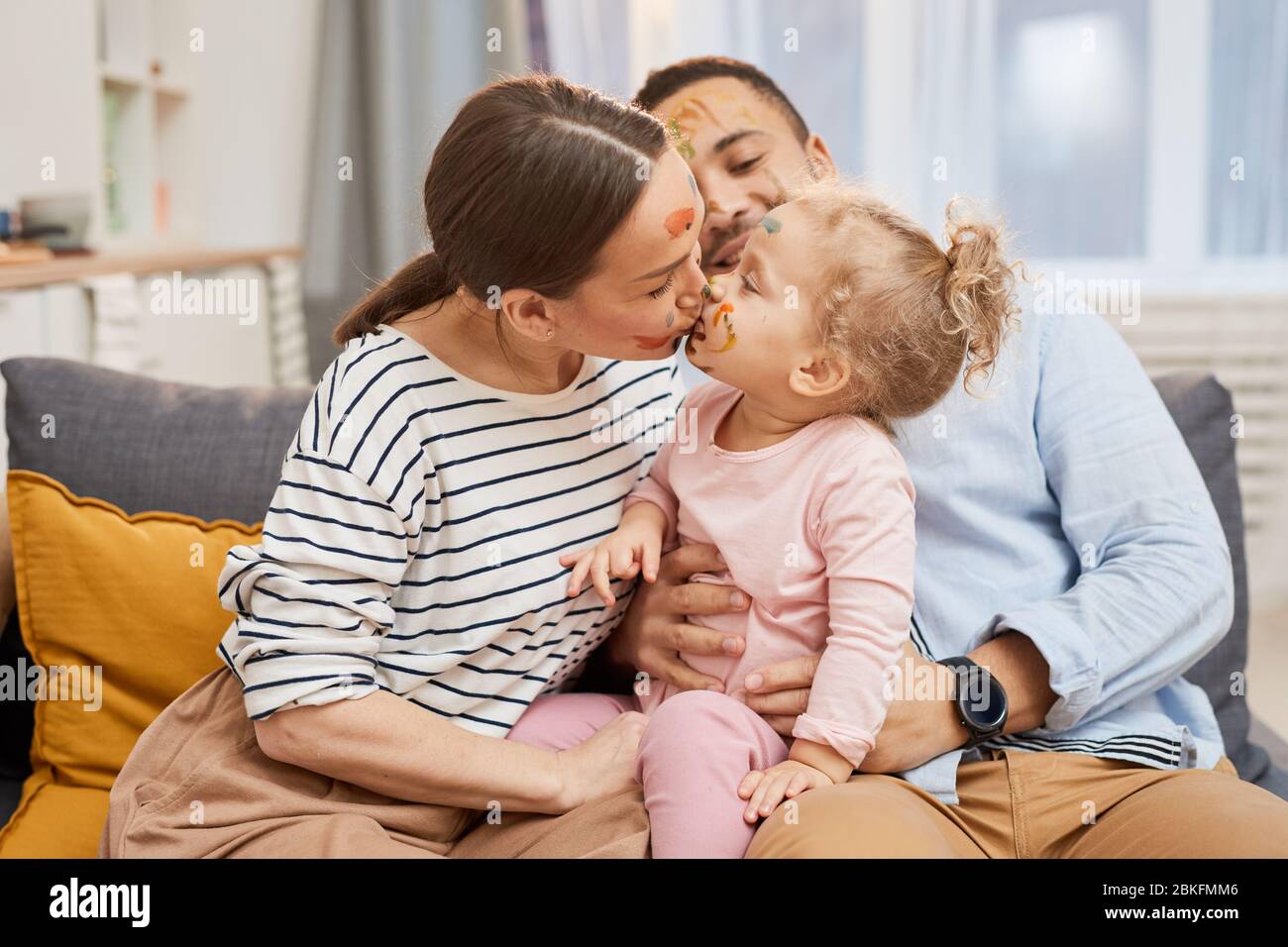 Glückliche junge Familie von drei zusammen auf dem Sofa sitzen, Mutter küsst ihre kleine Tochter, horizontale mittlere Aufnahme Stockfoto