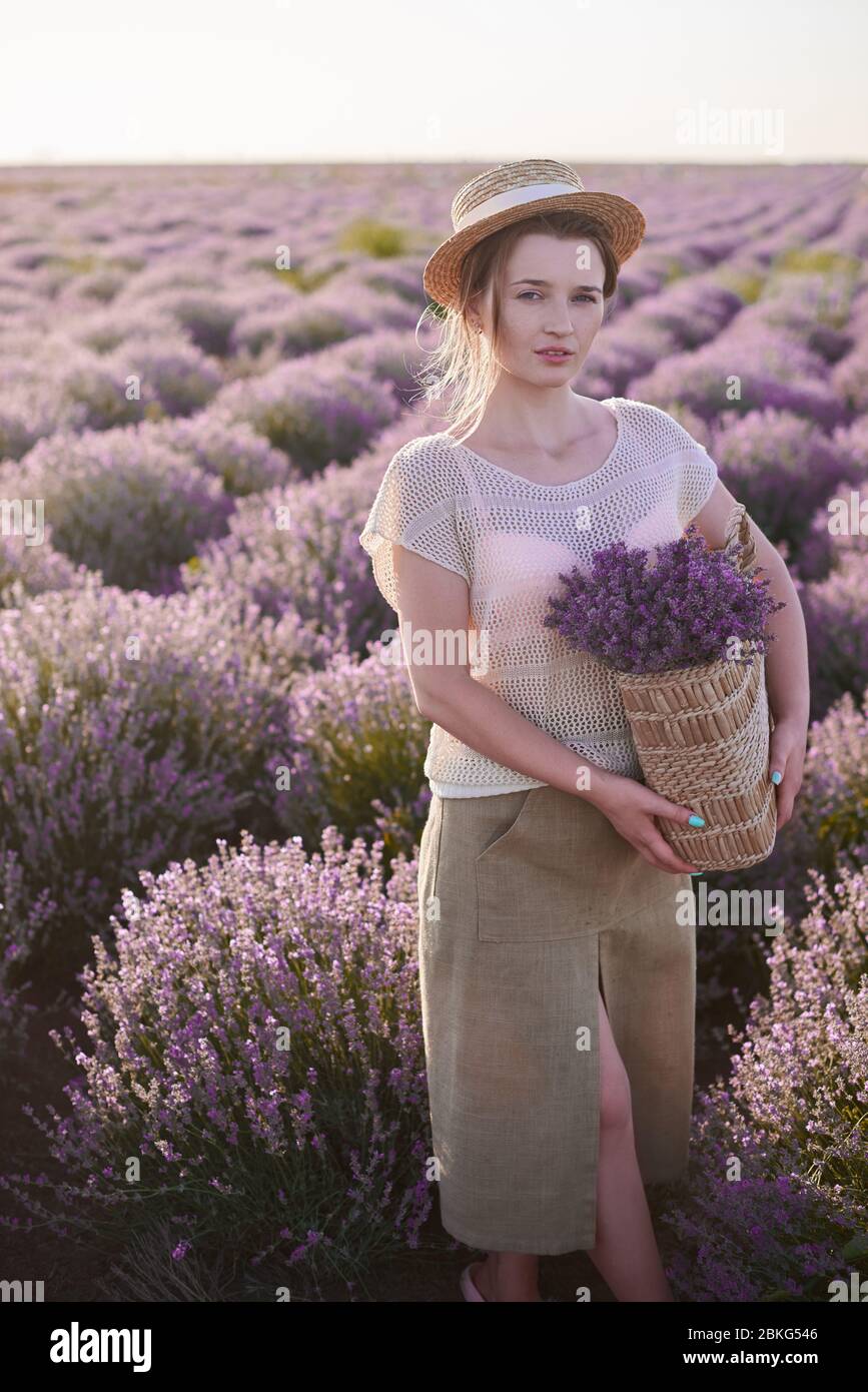 Junges Mädchen mit Weidenkorb mit Lavendel in den Händen in einem beigen Strohhut in den Lavendelfeldern Stockfoto