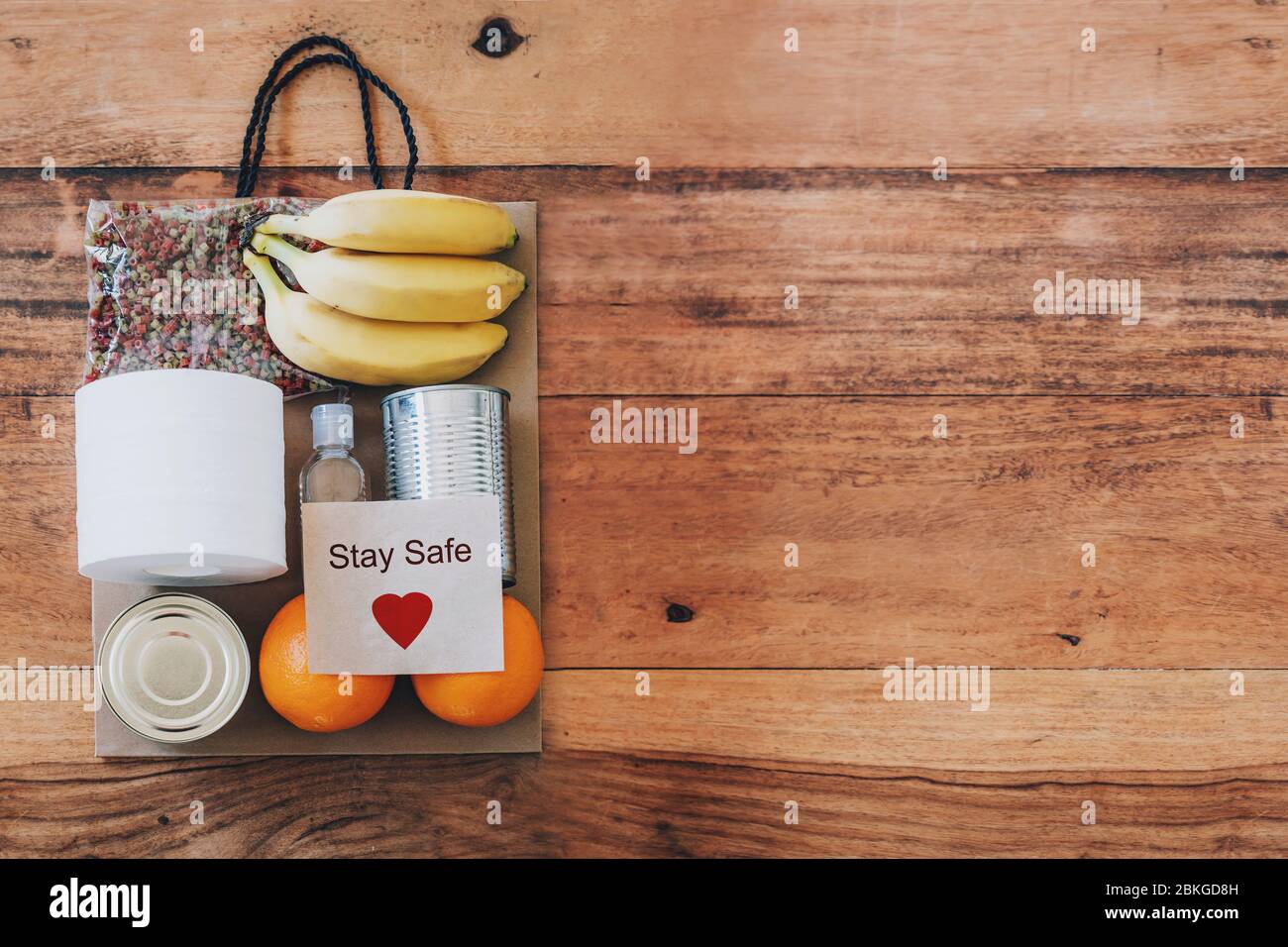 Papiertüte mit Lebensmitteln, Toilettenpapier, Handdesinfektionsmittel und Karte mit Schild Stay Safe darauf. Flach auf dem Holztisch liegen. Covid-19 Ausbruch, Selbstisolierung, Lieferung, Spendenkonzept. Stockfoto