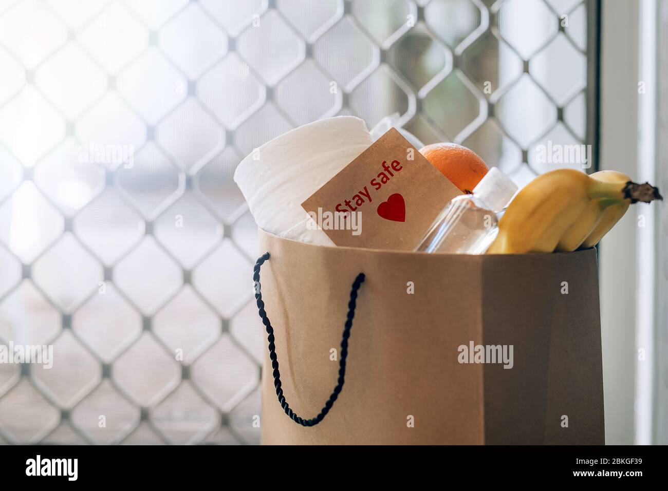 Papiertüte mit Lebensmitteln, Toilettenpapier, Handdesinfektionsmittel und Karte mit Schild Bleib sicher auf dem Boden am Eingang zum Haus. Covid-19 Ausbruch, Selbstisolierung, Lieferung, Spende. Stockfoto