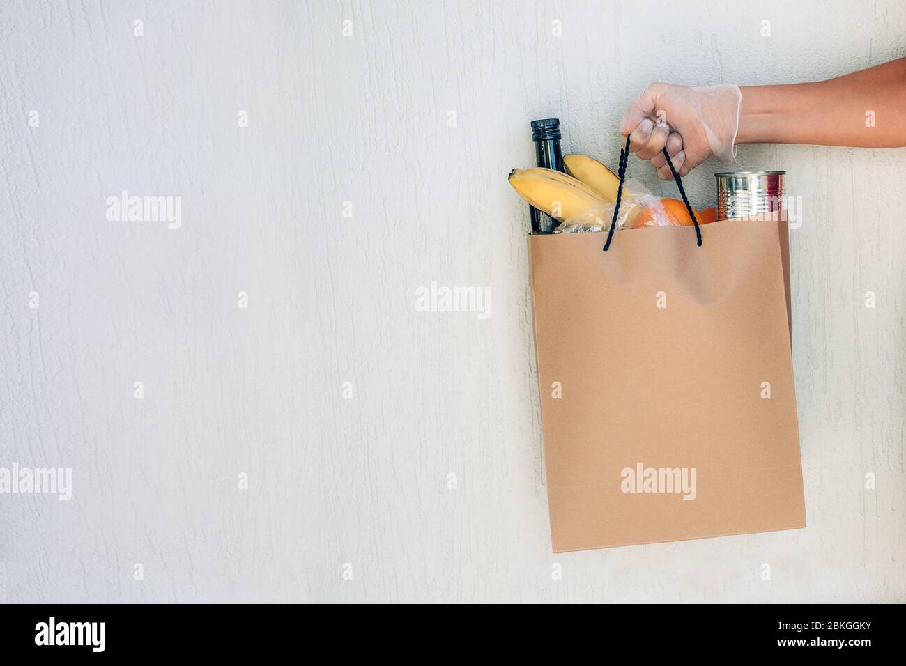 Papiertüte mit Lebensmitteln in der Hand der Frau mit Schutzhandschuh an der weißen Wand im Hintergrund. Covid-19 Ausbruch, Selbstisolierung, Lieferung, Spende. Stockfoto
