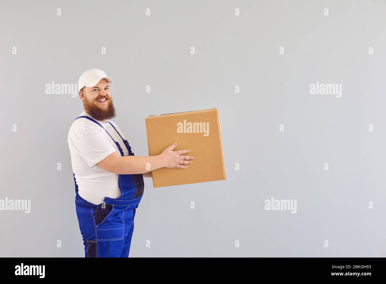 Lustige fette bärtige Lieferung Kurier Lader Mann in einer blauen Uniform mit einem Karton auf einem grauen Hintergrund. Stockfoto