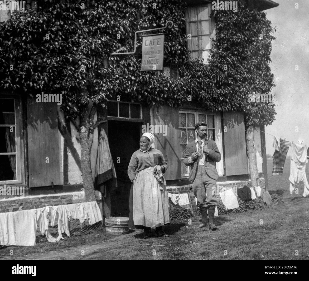 Ein französischer Mann und eine Frau stehen vor dem Cafe Amour in Marche-Neuf in der Normandie, Frankreich in den 1890er Jahren, mit all ihren Wäschestücke an den Linien. Stockfoto