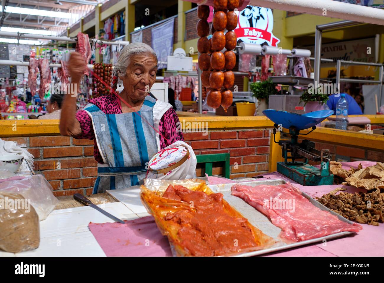 Indigene Dame mit Zöpfen, die einen Schal bestickt, während sie darauf wartet, dass Kunden an ihrem Fleischstand auf dem Zaachila-Markt ankommen. Stockfoto