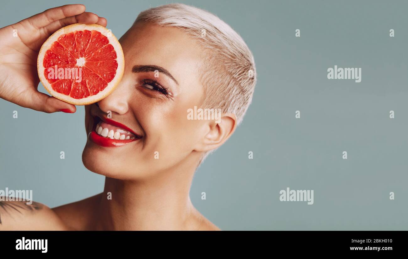 Nahaufnahme einer Frau, die vor grauem Hintergrund eine Grapefruit vor dem Auge hält. Schönes weibliches Modell mit kurzen blonden Haaren, die eine Grapefruit halten Stockfoto
