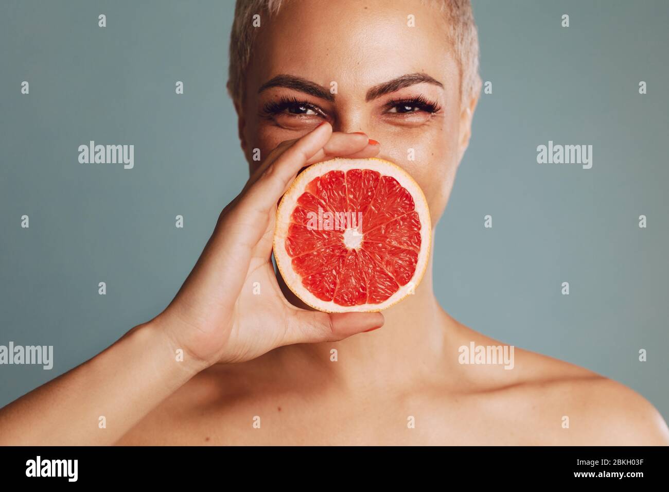 Nahaufnahme einer Frau, die vor grauem Hintergrund eine Grapefruit vor ihrem Mund hält. Schönes weibliches Modell mit einer Grapefruit-Scheibe. Stockfoto