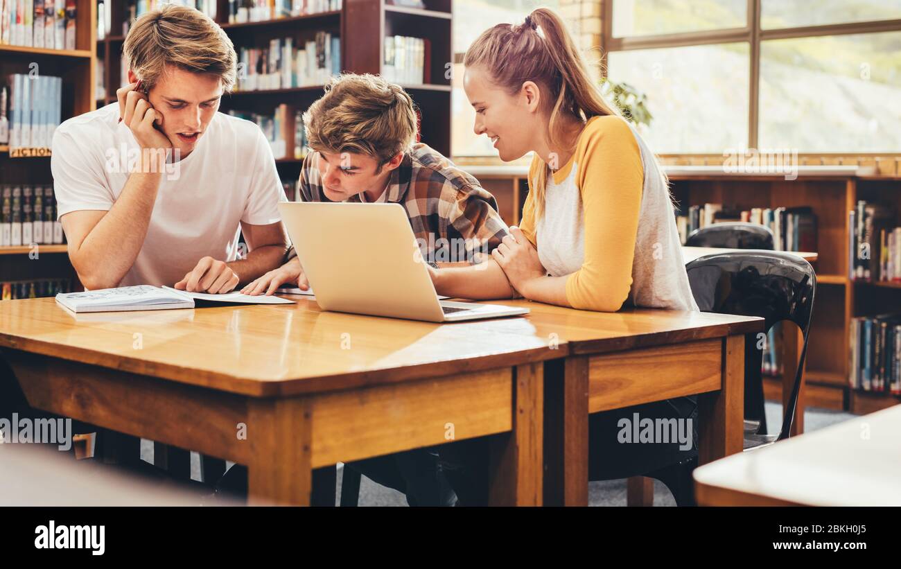 Schüler, die an Schulprojekt in der Bibliothek arbeiten, mit Laptop am Tisch sitzen und im Buch lesen. Junge Studenten, die in der Bibliothek studieren. Stockfoto