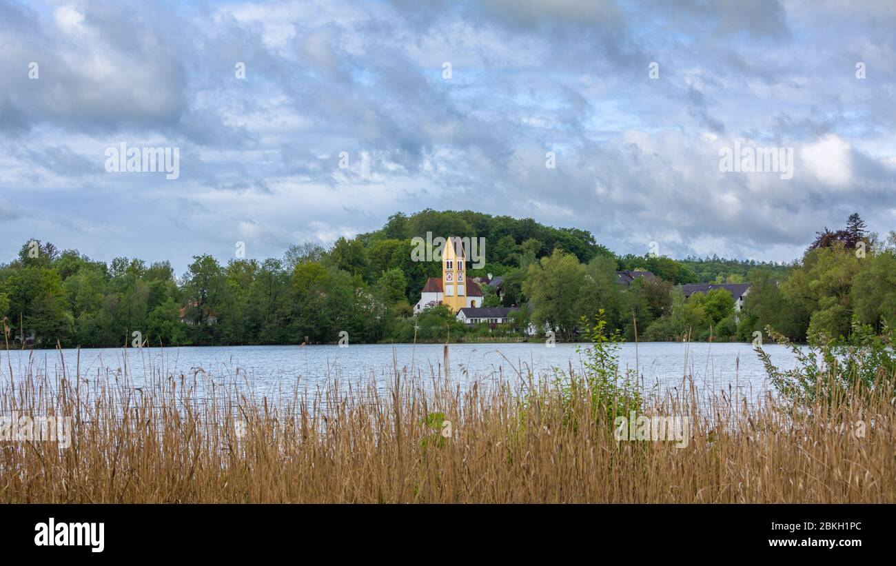 Wessling See mit Kirche 'Mariae Himmelfahrt' und üppigen, grünen Bäumen. Reed im Vordergrund. Perfekter Ort für Tagesausflüge von München. Panoramafarma. Stockfoto